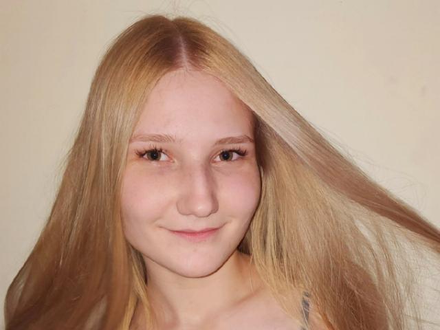 VanessaNorris has long blonde hair, light skin, small nose, subtle smile, and visible freckles. Her eyelashes are long and natural. She's wearing a thin-strapped top.