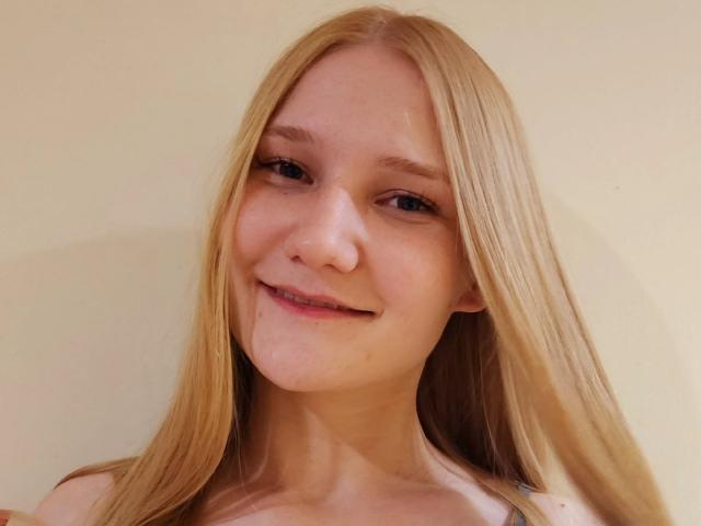 VanessaNorris has long blonde hair, fair skin, and a slight smile with a small drop of liquid on her chin. Her eyes are blue, and she's topless.