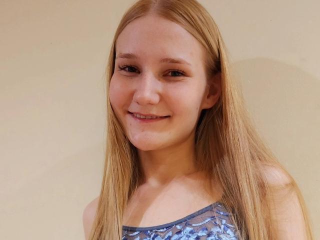 VanessaNorris has long, straight blonde hair, small tits, and a cute smile. She's wearing a blue, patterned, strapless top. Her skin is fair, and she's looking at the camera.
