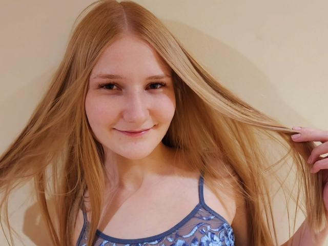 VanessaNorris with long blonde hair, smiling, holding her hair, wearing a blue lace top, showing her small tits, fair skin, cute face, and light makeup.