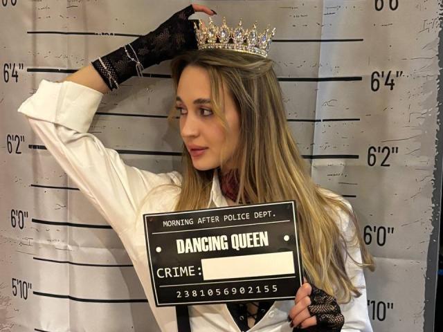 AlexaWilds, blonde, wearing a white police uniform, black lace glove, and silver crown, holding a sign with "Dancing Queen" and height, looking sideways, with a slight smirk.