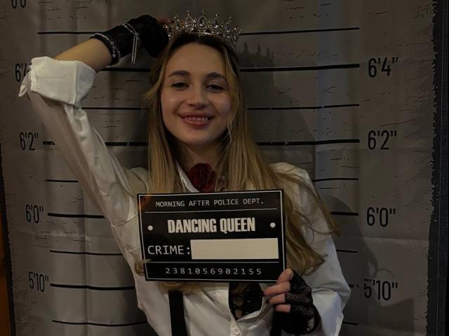 AlexaWilds, with long blonde hair, smiling, wearing a white shirt, black fingerless gloves, and a crown, holding a "Dancing Queen" sign, standing in front of a height chart.