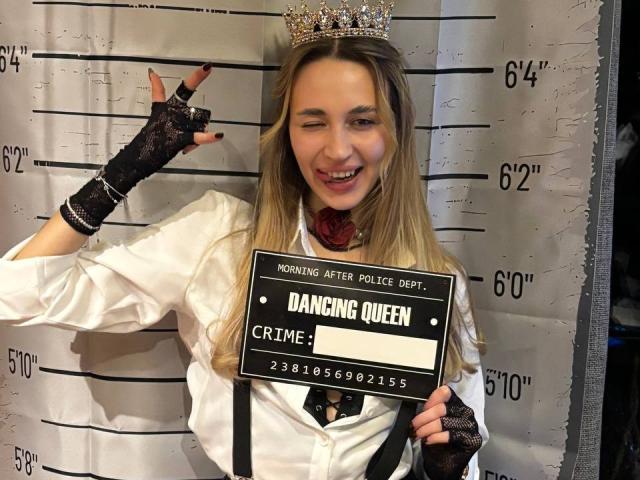 AlexaWilds with long blonde hair, wearing a silver crown, white shirt, black fingerless lace gloves, and a black necklace, smiling with one eye closed, showing a middle finger.