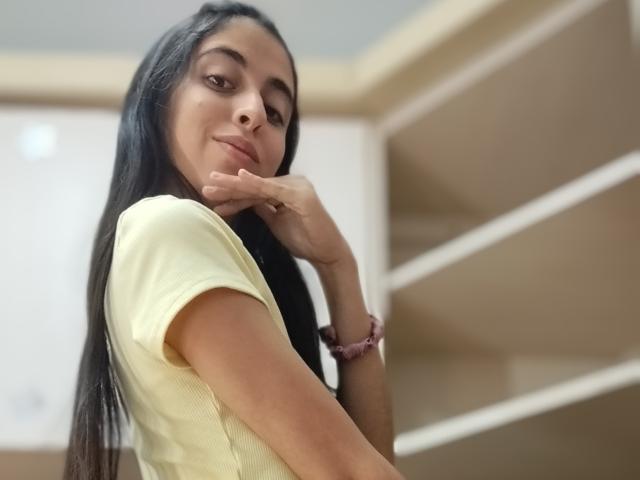 AnnaAlvarez with long black hair, yellow shirt, resting chin on hand, slight smile, brown eyes, bracelet on wrist, soft light, relaxed pose, looking down, subtle makeup, casual.