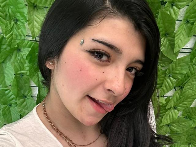 Close-up of SweetEescape with medium-length, straight black hair. She has light brown skin, brown eyes, and is sticking out her pink tongue slightly. She wears a white t-shirt and a gold chain necklace. A small, silver leaf-shaped sticker is on her left cheek. She has a few moles on her face and a subtle blush. Her eyebrows are dark and well-defined. The green leafy background contrasts with her skin tone. The image is sharp and well-lit.