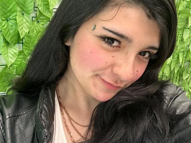 SweetEscape has black hair, brown eyes, and a cute green leaf on her cheek. She's smiling with pink blushed skin, wearing a black leather jacket and brown necklace.