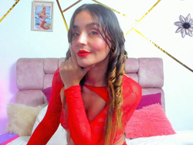 MindyLove with long braided hair, red lipstick, and red mesh top, touching her cheek, showing her red cleavage, sitting on a pink bed with fluffy pillows.
