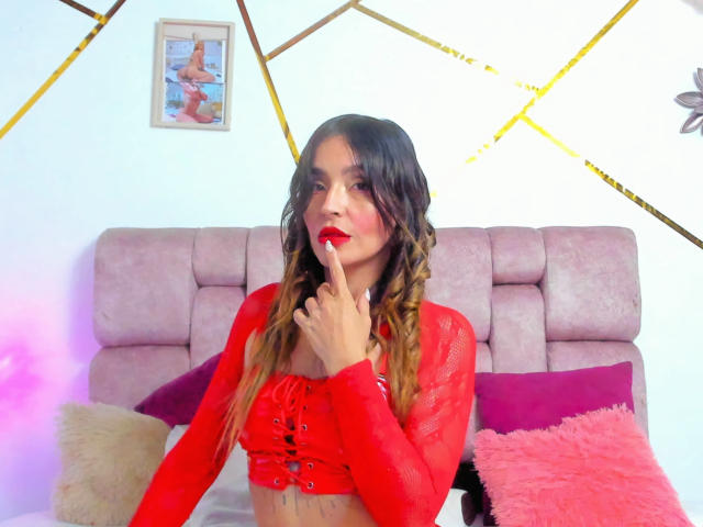 MindyLove with red mesh top, finger in mouth, red lipstick, braided brown hair, sitting on pink bed, pink and purple pillows, headboard in background, framed picture on wall.