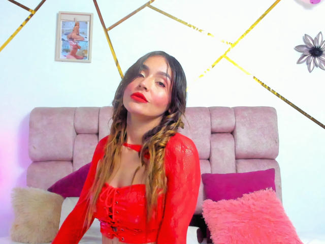 MindyLove with long, wavy brown hair wearing a red lace-up corset, sitting on a pink bed, red lipstick, and a confident, slightly playful expression. Boobs, cute, sexy.