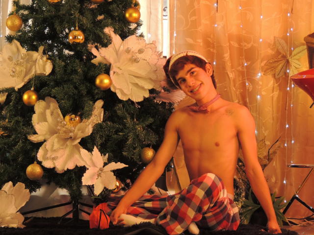 JacobeLord, shirtless, sitting in front of a Christmas tree with large white poinsettia decorations and gold ornaments. He wears a pink choker and plaid pajamas, with a red cap tilted on his head. His skin is light, and he has short brown hair. He's smiling slightly, with one hand resting on the floor and the other on his knee. The background has warm, orange curtains with string lights. The scene is cozy and festive.