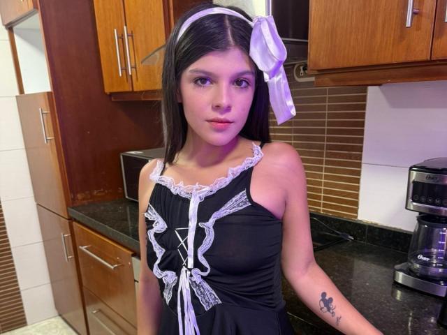 RilleyFoyx with dark hair, white headband, purple bow, black lace-up dress, small tits, tattoo on right arm, standing in kitchen.