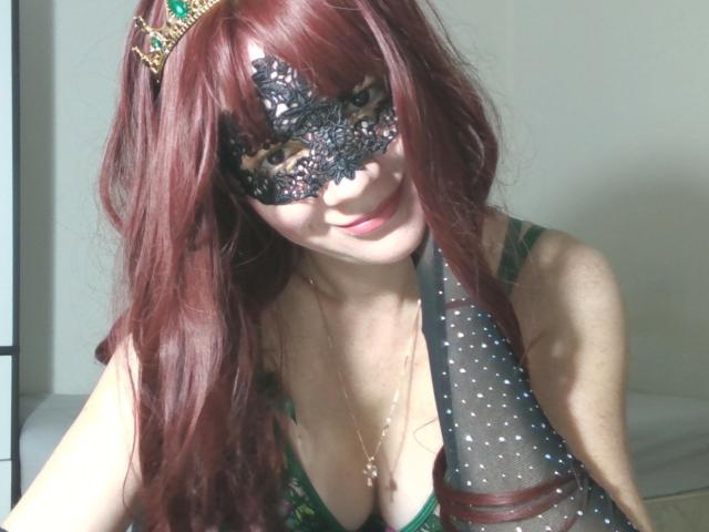 Aromalove with long red hair wears a black lace mask and a green, sheer polka-dot dress. She has a small smile and a gold necklace with a cross pendant. A green and gold tiara is on her head. Her skin is light, and she's sitting in a brightly lit room. The image has a casual, slightly overexposed look. Her cleavage is visible, and the focus is on her upper body.
