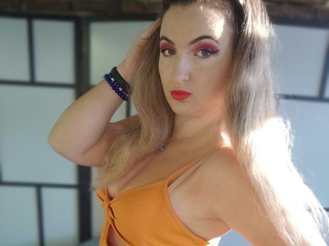 GoddessMonicaAngels with red lipstick, orange ribbed top, blonde hair, pink eyeshadow, blue bracelet, hand in hair, sunlight, visible tits, serious expression.