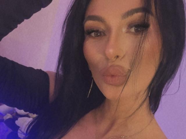 KinkyAmelie with pouty lips, dark long hair, thick eyebrows, eyeliner, and nose ring, wearing a black sleeve and long earrings, making a kissing face.