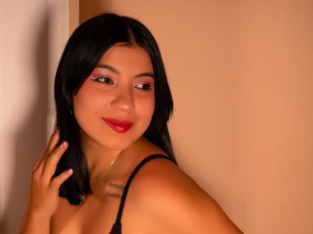 AveryTorres with dark hair, pink eyeshadow, red lipstick, black strap, smiling, hand on shoulder, small tattoo, tan skin, gold necklace, against beige wall.