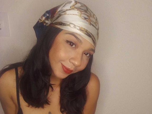 NanaMilan with dark hair, red lipstick, and a white patterned headscarf, smiling softly. She's wearing a black spaghetti strap top, showing a small tattoo on her chest.