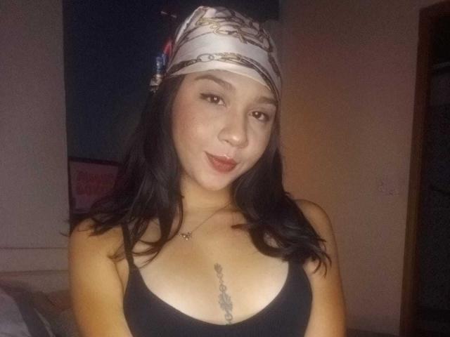 NanaMilan with black hair, wearing a headscarf, black tank top, and necklace, showing small tits with a tattoo under her chest, smiling at the camera.