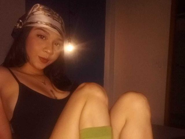 NanaMilan with dark hair, wearing a headband, black tank top, and green socks, sitting with knees up, smiling at camera, light in background, showing small boobs and ass.