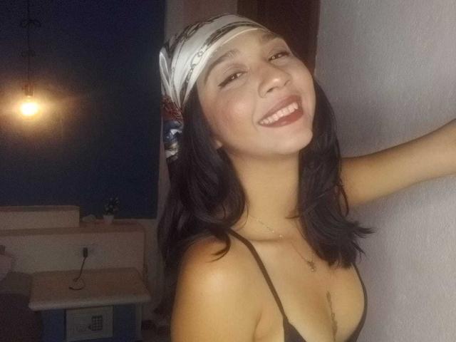 NanaMilan tem cabelo escuro, uma faixa de cabeça branca e um top de biquíni preto, sorrindo com o braço levantado. Ela está mostrando os seios e tem um rosto fofo e feliz..