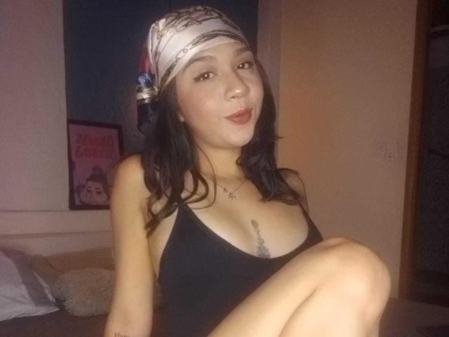 NanaMilan mit dunklem Haar, die ein weißes Kopftuch trägt, ein schwarzes Tanktop, das ihre Brüste zeigt, sitzt mit verschränkten Beinen, lächelt, trägt roten Lippenstift, hat ein Tattoo auf der Brust, im Schlafzimmer..