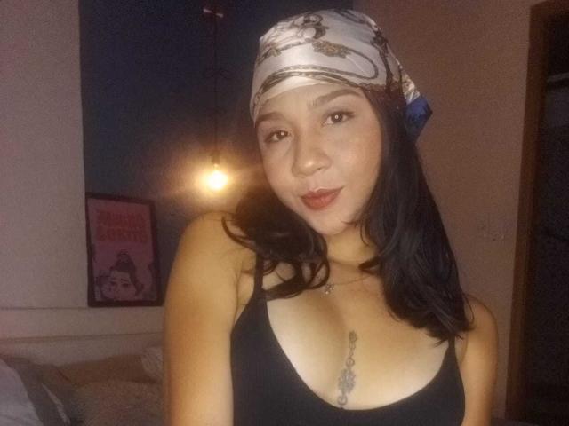 NanaMilan, avec ses cheveux foncés, portant un foulard blanc et gris, un débardeur noir laissant entrevoir de petites seins, un tatouage sous les seins, peau claire, souriante, dans une pièce peu éclairée..
