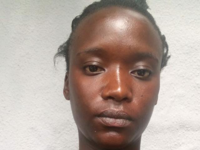 Close-up of BlackFirre with dark brown skin, small black pupils, and subtle makeup. BlackFirre has thin, curly hair pulled back, a slightly shiny forehead, and full, slightly pressed lips. The texture of BlackFirre's skin is smooth with a few small, natural blemishes. The face is centered against a plain, textured white background. BlackFirre's expression is neutral, looking slightly to the right.