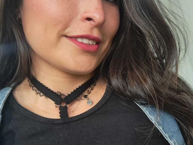AlexandraSaenz69's face with pink lipstick, slight smile, black lace choker, black shirt, denim jacket, and a small pendant, showing her cute nose and partial brown wavy hair.