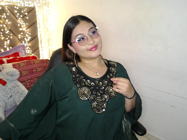 HelueJamil with glasses, pink lipstick, and green dress with black floral embroidery, smiling, hand on chest, white wall behind, chair, and string lights.