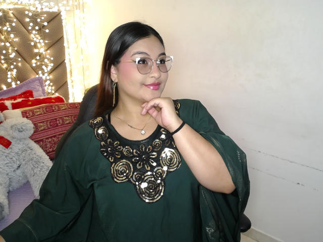 HelueJamil with glasses, pink lipstick, green dress, shiny black and gold floral chest, black hair, right hand near chin, white wall, string lights, and a fluffy gray toy.