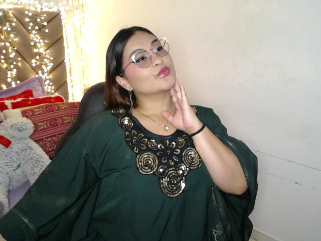 HelueJamil with glasses, pink lipstick, green dress, ornate black and gold chest piece, hand on cheek, black hair, small breasts, fair skin, slightly chubby, white wall background.