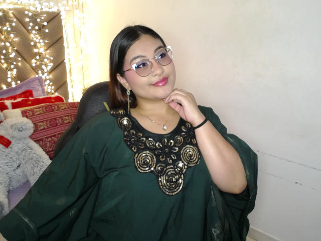 HelueJamil, chubby Asian woman with glasses, dark green top, black floral necklace, pink lipstick, smiling, hand near face, black hair, white wall, string lights in background.
