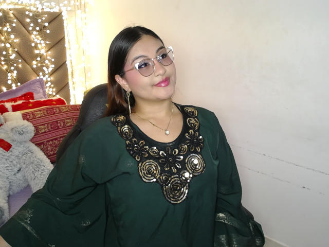 HelueJamil, a chubby woman with dark hair, glasses, and pink lipstick, wearing a green top with ornate gold rose patterns, smiling in a room with string lights.