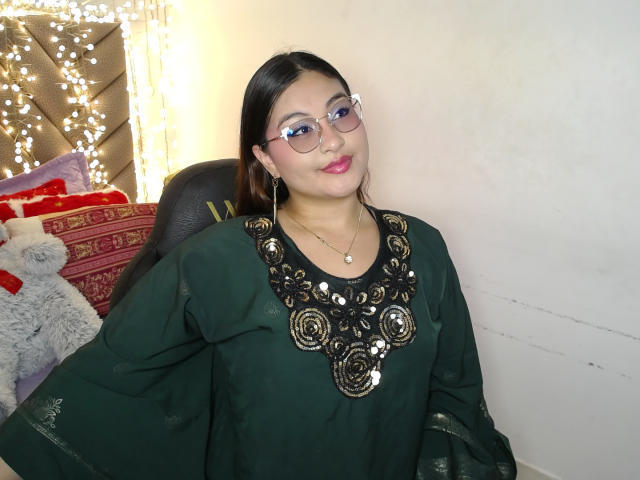 HelueJamil, Asian woman with glasses, green dress with black and gold embroidery, small tits, pink lipstick, smiling, white wall, black chair, stuffed animal, and string lights in background.