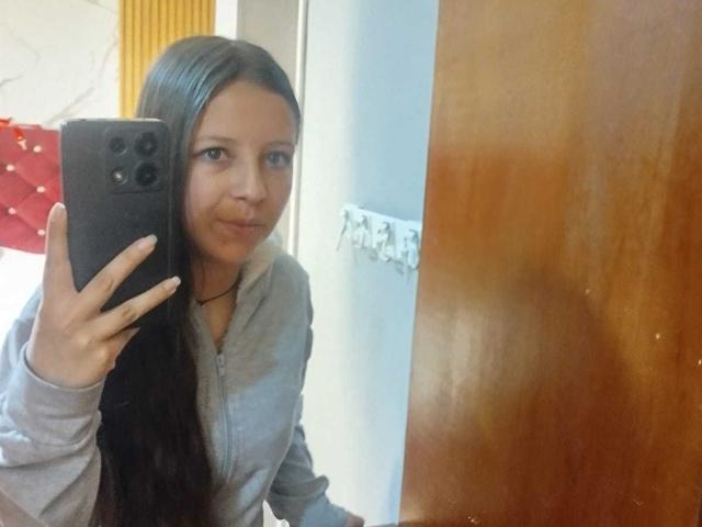 GraceRedMiller with long dark hair, pouty face, holding black phone, wearing gray hoodie, taking mirror selfie, wooden door on right, white wall with hook in background.