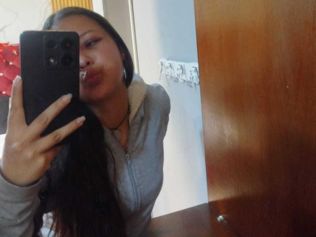 GraceRedMiller, with long black hair, pouting lips, and wearing a grey hoodie, takes a mirror selfie with a black phone, showing her chest in the reflection.