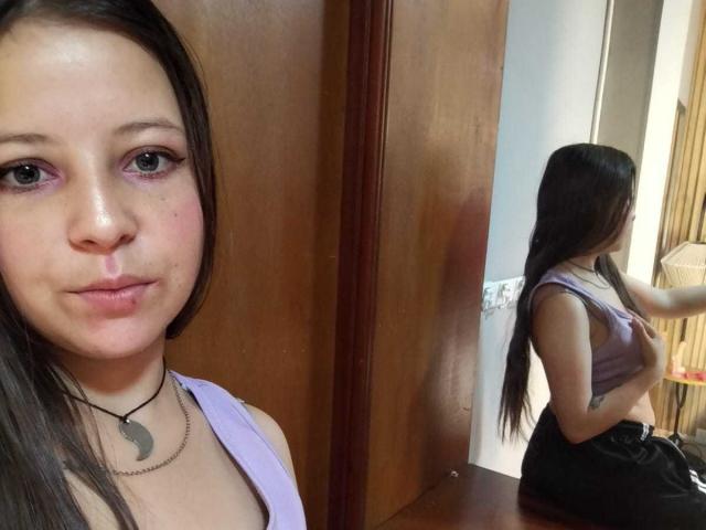 GraceRedMiller with long brown hair, purple eye makeup, and a yin-yang necklace, showing her small tits in a purple top, reflected in a mirror, standing in front of wood.