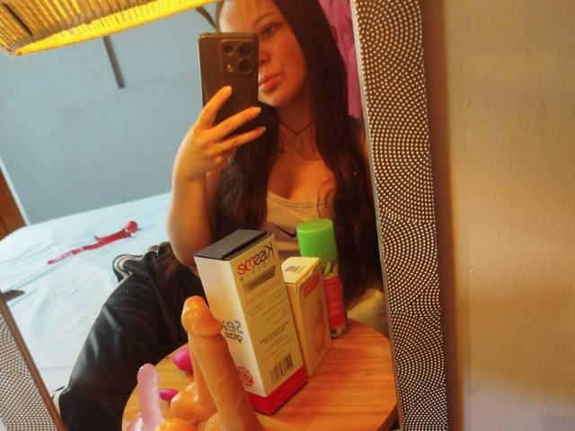 GraceRedMiller with long dark hair, taking a mirror selfie, wearing a low-cut top showing her tits, holding a dildo and sex toys on a wooden table.