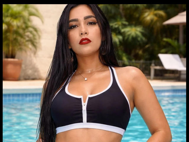 DarshaJaya, con lunghi capelli neri, rossetto rosso e un reggiseno sportivo nero con bordi bianchi, che mostra il seno, è in piedi accanto a una piscina blu. Indossa una collana con un piccolo ciondolo..