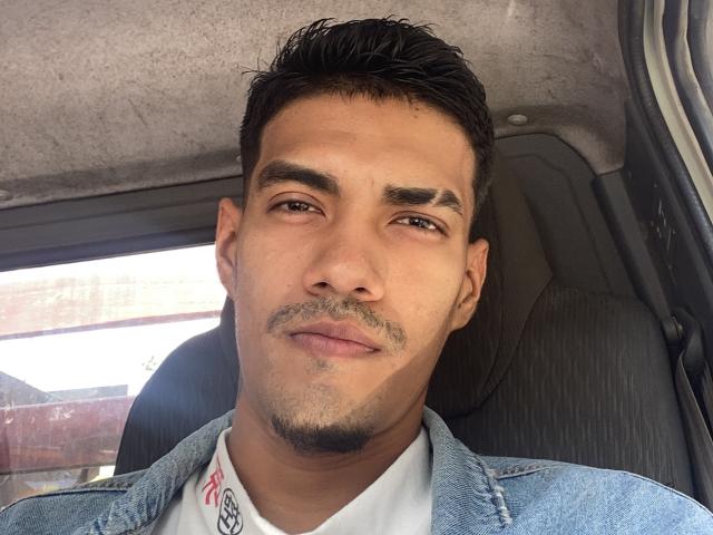 MikaelGrey has a medium skin tone, short dark hair, and a light mustache. He's wearing a denim jacket over a white shirt, sitting in a car.