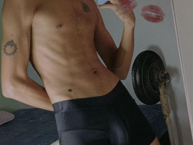 MikaelGrey, shirtless, wearing black boxer briefs, showing a slim, toned, hairy chest, sun tattoo on left arm, holding phone, standing in front of a fan, green wall.