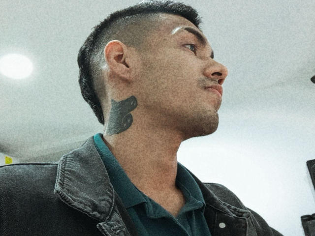 MikaelGrey has a black tattoo of a bird on his neck, short dark hair, and a nose ring, wearing a green shirt and black jacket, looking to the side.