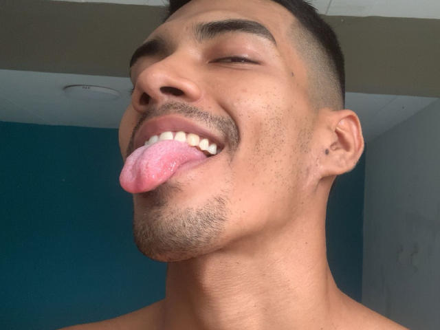 MikaelGrey's face with short dark hair, brown skin, and a goatee, sticking out his pink tongue, eyes closed, and a small tattoo on his right ear.