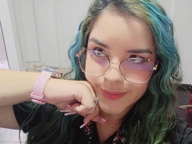 AuroraSantorini with green-blue hair, pink glasses, purple eyeshadow, pink wristwatch, and pink nail polish, resting her chin on her hand, smiling. Boobs not visible.