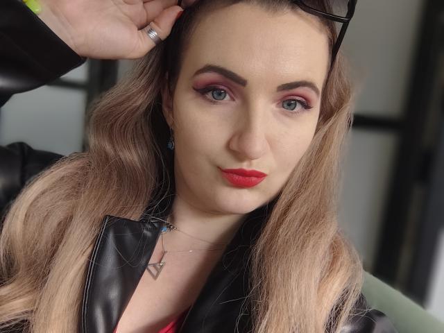 GoddessMonicaAngels with red lipstick, pink eyeshadow, and black leather jacket, holding sunglasses, long blonde hair, blue eyes, and a silver necklace with a triangular pendant.