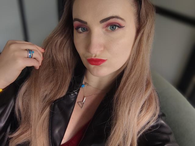 GoddessMonicaAngels has blonde hair, red lipstick, pink eyeshadow, blue ring, black leather jacket, and a necklace with a blue gem. She's looking at the camera, slightly pouting.