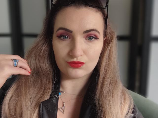 GoddessMonicaAngels with red lipstick, pink eyeshadow, and blue eyes, wearing black sunglasses on her head, a black leather jacket, and a blue ring, holding her hair.
