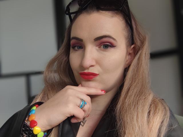GoddessMonicaAngels with red lipstick, pink eyeshadow, and blonde hair, touching her chin with a finger, wearing colorful candy bracelet, black leather jacket, and sunglasses on head.