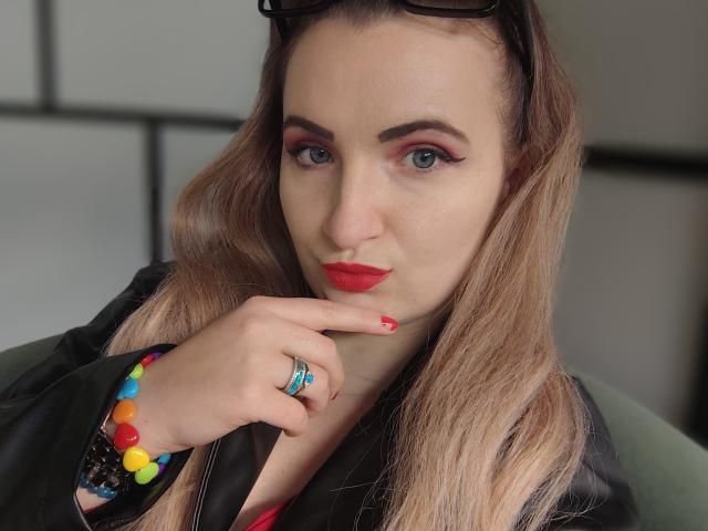 GoddessMonicaAngels with red lipstick, finger to chin, black jacket, colorful beaded bracelet, blue ring, red nails, eyeglasses on head, light brown hair, and pink eyeshadow.