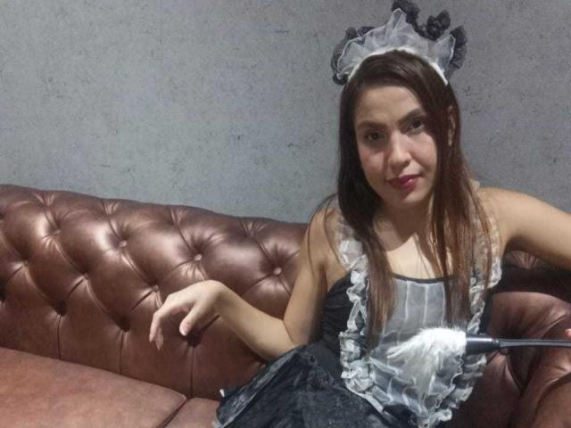SusanThomass with long brown hair, wearing a black and white maid outfit, sitting on a brown tufted leather couch, holding a feather duster. Her tits are visible.