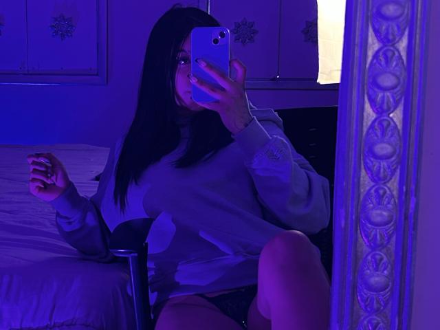 KatianPierce with dark hair, wearing a loose gray sweater, taking a mirror selfie in purple-blue lighting, sitting on a chair with a bed behind her. Visible ass.